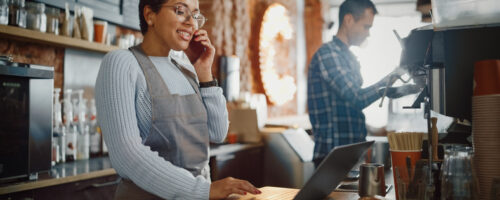 Latin American Coffee Shop Employee Accepts a Pre-Order on a Mobile Phone Call and Writes it Down on Laptop Computer in a Cozy Cafe. Restaurant Manager Browsing Internet and Talking on Smartphone.