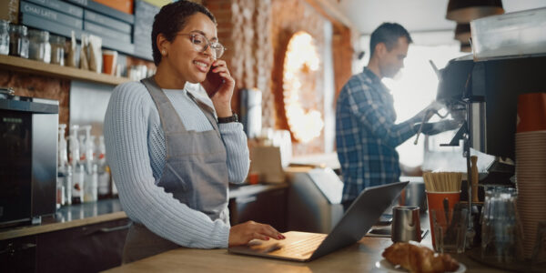 Latin American Coffee Shop Employee Accepts a Pre-Order on a Mobile Phone Call and Writes it Down on Laptop Computer in a Cozy Cafe. Restaurant Manager Browsing Internet and Talking on Smartphone.